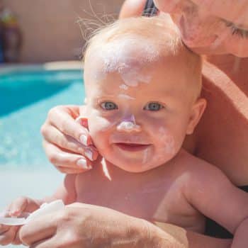 A mother putting sun screen on a 10 month old baby. The mother is rubbing sun screen into the baby boys face in a back yard. A swimming pool can be seen in the background.