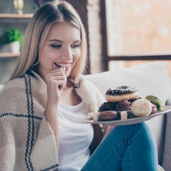 Portrait of beautiful emotional charming attractive sweet toothy woman sitting on sofa in living room, holding plate of donuts and macaroons, looking exciting satisfied (Portrait of beautiful emotional charming attractive sweet toothy woman sitting on