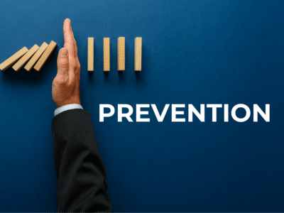 A person wearing a suit uses their hand to stop a line of falling wooden dominoes, preventing the remaining upright dominoes from toppling. The word 'PREVENTION' is displayed in bold white text to the right of the hand, set against a dark blue background