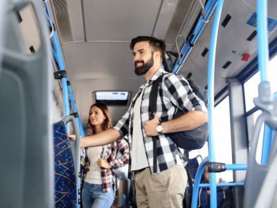 A young man with a beard and backpack stands inside a bus, holding onto a railing, while a woman in the background, also wearing a backpack, walks down the aisle. Both are dressed casually in plaid shirts, appearing comfortable during their commute. The bus interior features blue rails and patterned seats, creating a typical public transportation setting