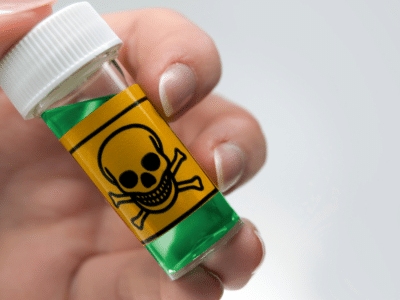 A close-up of a hand holding a small glass vial containing green liquid. The vial is labeled with a yellow warning sticker displaying a skull and crossbones, indicating that the substance inside is toxic or hazardous