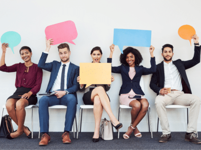 A diverse group of five professionals sit in a row on chairs, each holding up colorful speech bubbles above their heads. They are dressed in business attire, smiling, and engaging, with various expressions. The speech bubbles are in different colors: blue, pink, green, yellow, and orange. One person holds a coffee cup, while others hold notebooks or tablets, creating a lively and collaborative atmosphere.