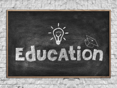 A chalkboard with the word 'Education' written in large, bold, white chalk letters. Above the letter 'A' is a drawing of a lightbulb, symbolizing an idea, and to the right is a graduation cap, representing learning. The chalkboard is framed with wood and set against a white brick wall background