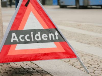 A red triangular warning sign on the street with the word 'Accident' displayed prominently in white text on a black stripe. The sign is set on a cobblestone surface, with a blurred background of the road and a passing vehicle, indicating an accident or caution zone