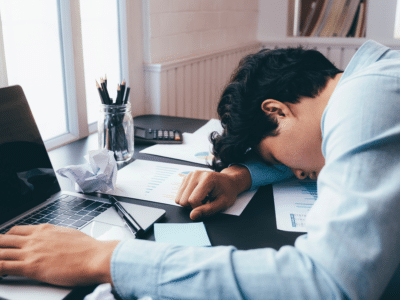 A man in a light blue shirt is slumped over his desk, asleep with his head resting on documents displaying charts and graphs. A laptop, crumpled paper, a pen, and a calculator are scattered across the desk, indicating exhaustion or overwork. The scene is set near a window with natural light coming in